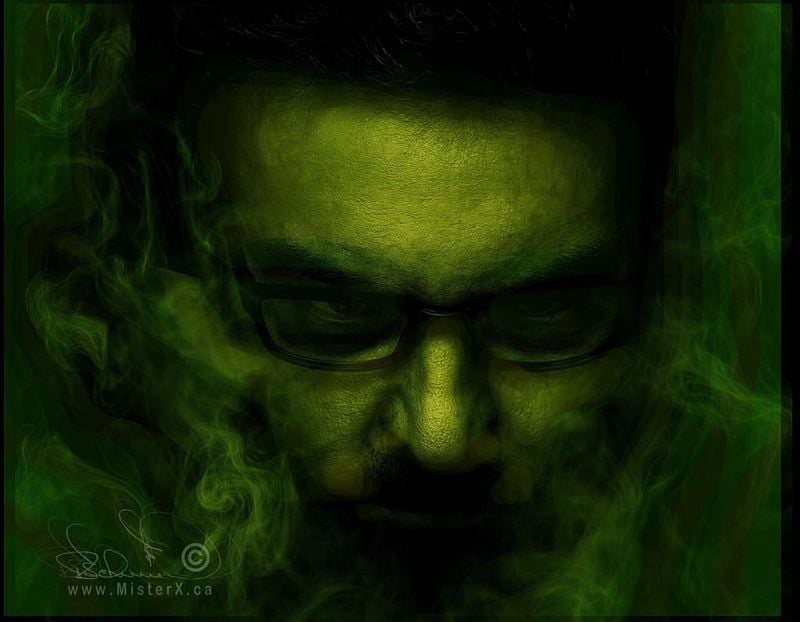 This image features a dark, moody portrait of a man's face cast in an intense green glow, emerging from a heavy shroud of wispy green smoke or vapor. He is looking downward with a serious, intense expression, wearing dark-rimmed glasses that obscure his eyes in shadow. The lighting strongly accentuates the texture of his skin and the bridge of his nose, while the rest of his head blends into a pitch-black background.