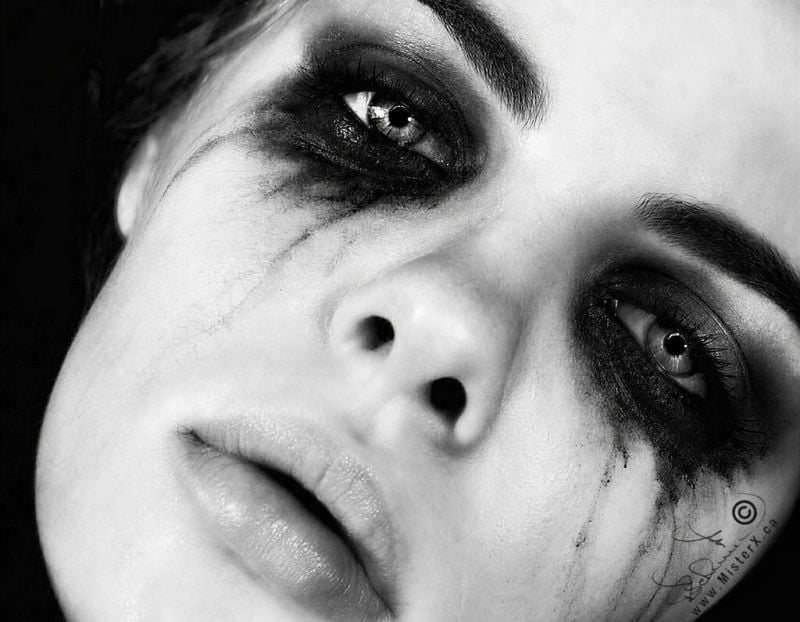A close-up, high-contrast black and white photograph captures the tilted face of a person with heavy, smeared dark makeup surrounding their eyes. The intense, dark eyeshadow is smudged unevenly around the eye sockets, with fine streaks resembling tears running down the cheeks. The individual's eyes are wide open, reflecting a sharp light source, while their lips are slightly parted in a neutral or somber expression. The background is entirely black, drawing full focus to the textured details of the skin and the dramatic makeup. A stylized signature and copyright symbol alongside the text "www.MisterX.ca" is visible in the lower right corner of the frame.