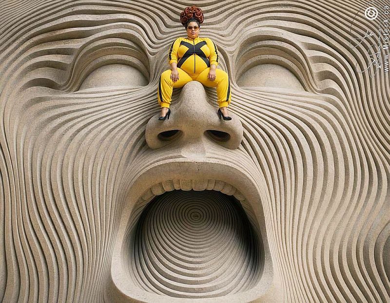 A plus sized woman in yellow tracksuit sits upon the nose of a detailed psychedelic stone sculptured screaming face.